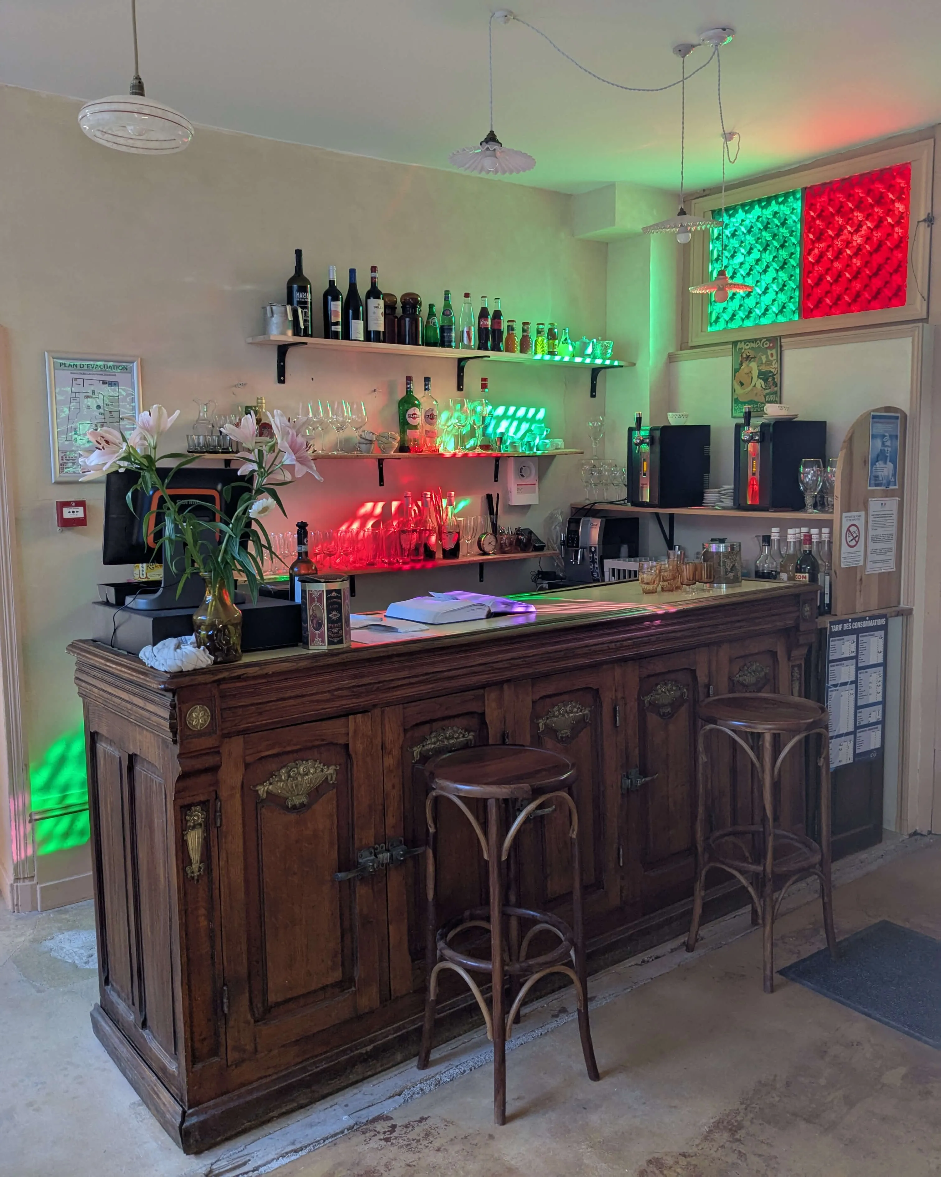 Accueil d'un restaurant avec un comptoir en bois ancien et des tabourets hauts. Derrière le bar, des bouteilles de vin et de champagne sont disposées. Ambiance chaleureuse, sous des vitraux colorés apportant une touche rétro.