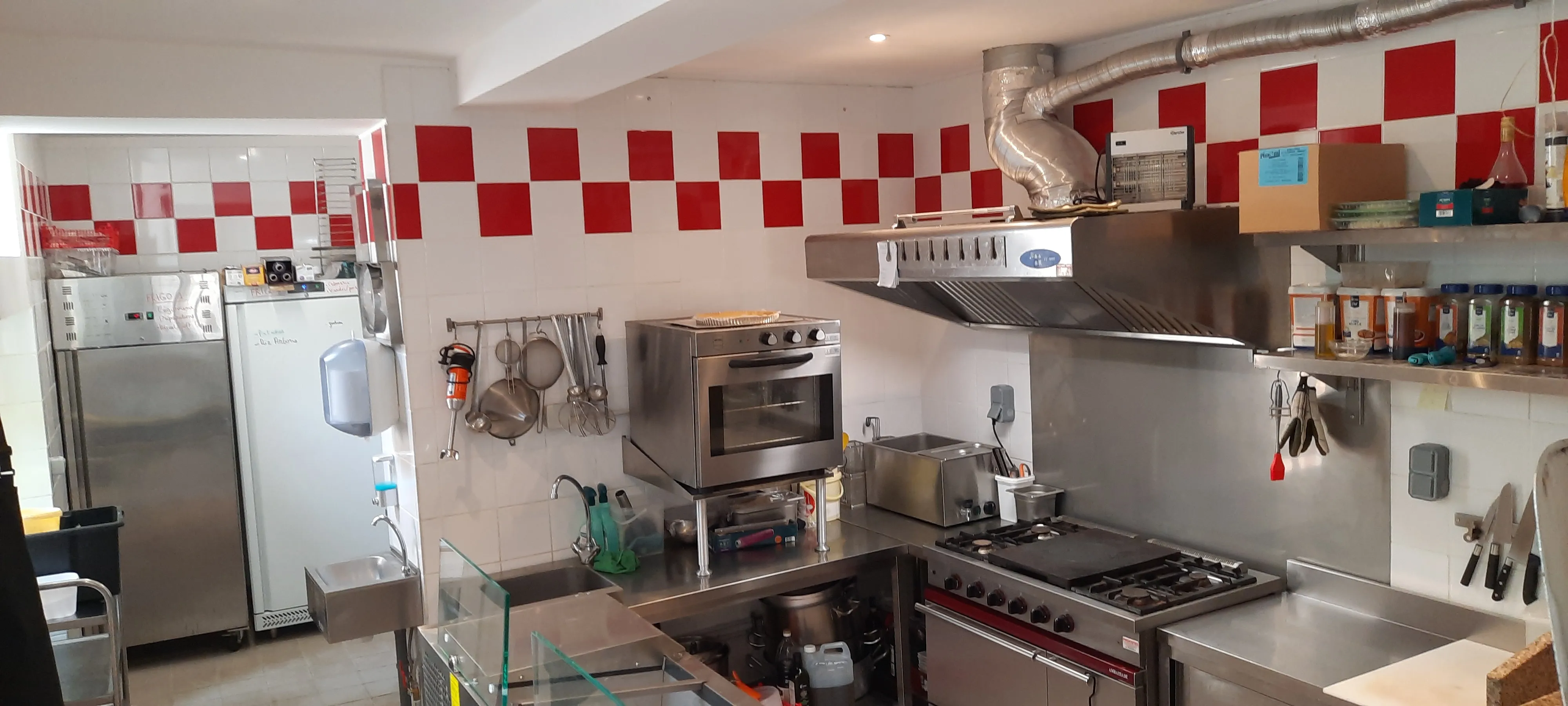 Cuisine professionnelle de restaurant avec équipements en inox, four professionnel, plaques de cuisson à gaz, hotte aspirante, étagères d'épices, ustensiles suspendus et carrelage mural rouge et blanc – cuisine équipée conforme aux normes HACCP