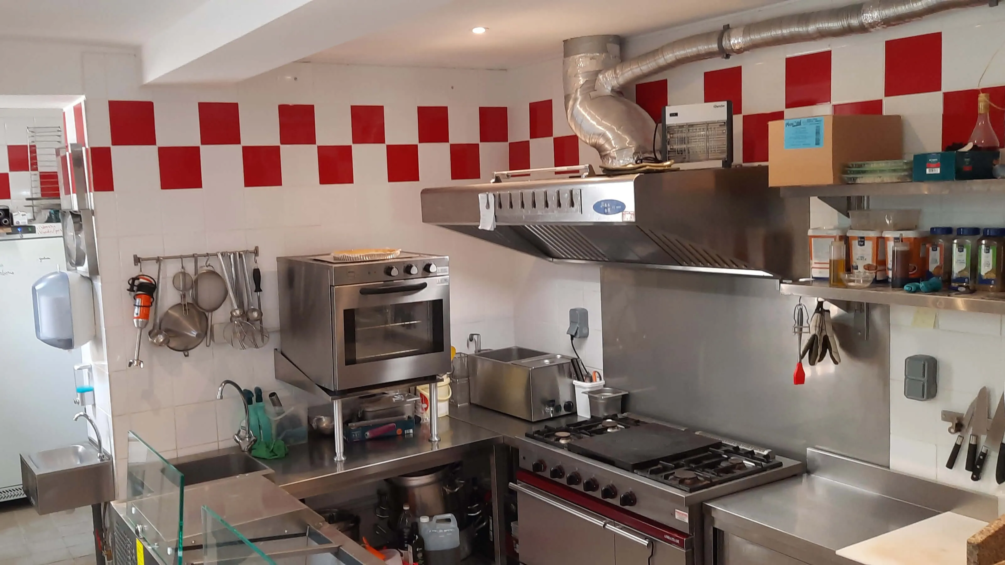 Cuisine professionnelle de restaurant avec équipements en inox, four professionnel, plaques de cuisson à gaz, hotte aspirante, étagères d'épices, ustensiles suspendus et carrelage mural rouge et blanc – cuisine équipée conforme aux normes HACCP