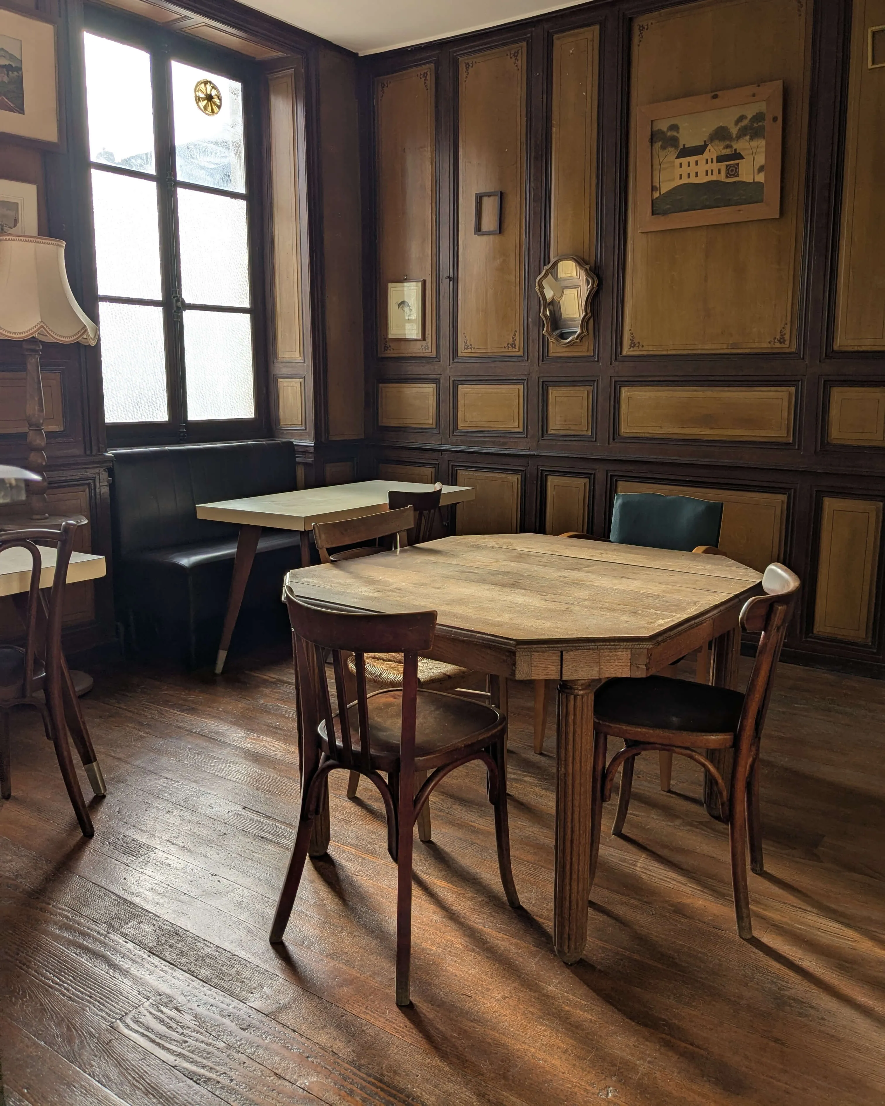 Salle de restaurant chaleureuse avec boiseries anciennes, cheminée et fauteuils verts vintage. Des clients dégustent un repas dans une ambiance conviviale et authentique.