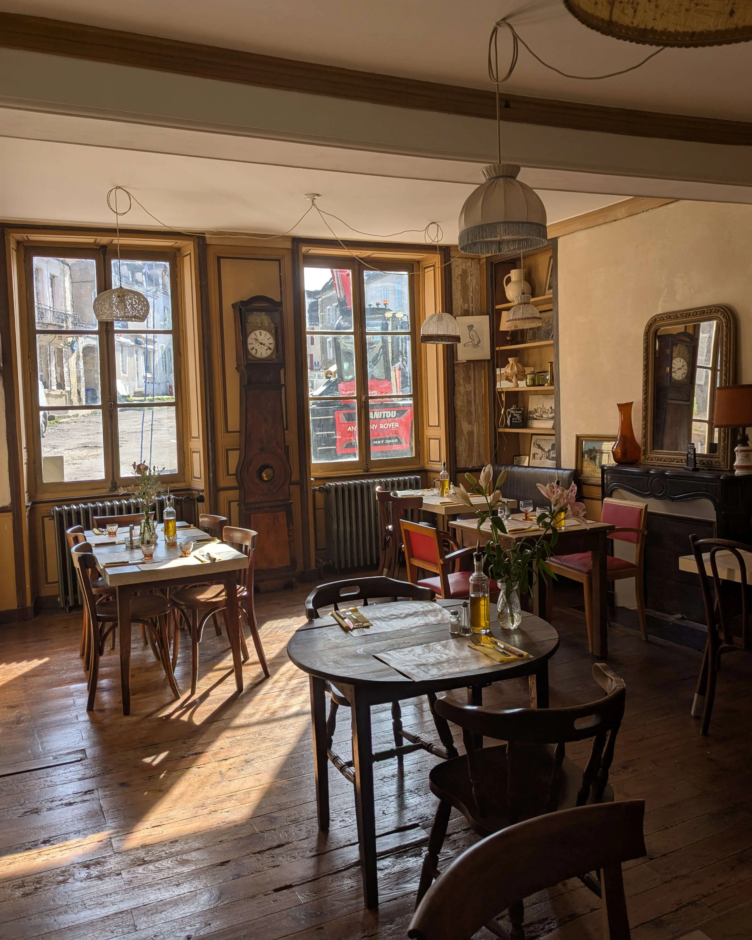 Salle de restaurant chaleureuse et lumineuse avec parquet ancien, tables en bois dressées, décoration vintage et grandes fenêtres donnant sur une place de village.