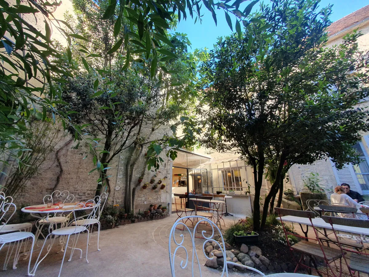 Vue depuis l’intérieur d’un restaurant sur une cour calme et arborée, vieux murs en pierre et ambiance rustique.